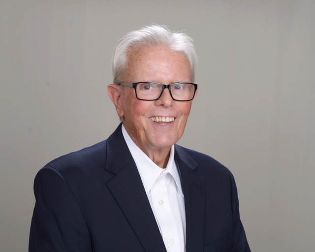 A man in glasses and a suit standing next to a wall.